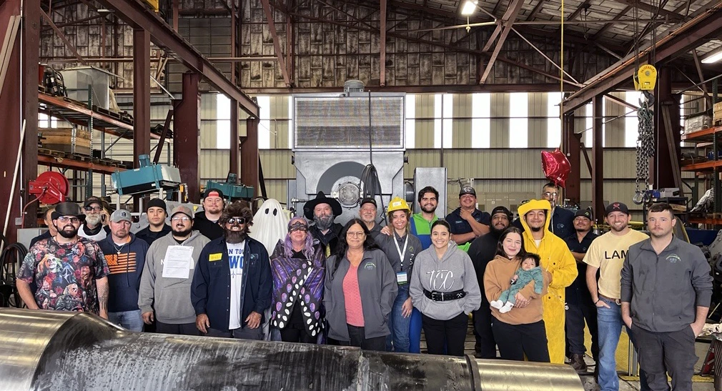 Group of ABS Wind employees wearing Halloween costumes and posing for a photo inside the Big Spring, Texas workshop, surrounded by industrial equipment and gearbox components.